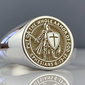 May include: A silver ring with a circular emblem depicting a knight with a sword and shield bearing a cross. The ring is engraved with the text "PUT ON THE WHOLE ARMOR OF GOD" and "EPHESIANS 6:13-17".