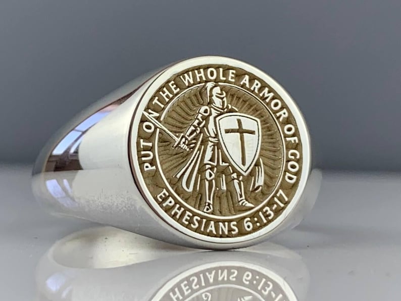 May include: A silver ring with a circular emblem depicting a knight with a sword and shield bearing a cross. The ring is engraved with the text "PUT ON THE WHOLE ARMOR OF GOD" and "EPHESIANS 6:13-17".