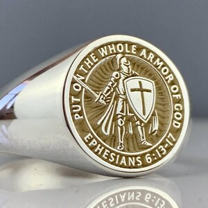 May include: A silver ring with a detailed circular emblem. The emblem features a knight with a sword and shield, surrounded by the text "PUT ON THE WHOLE ARMOR OF GOD" and "EPHESIANS 6:13-17". The ring has a polished, reflective surface.