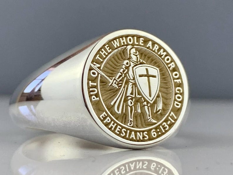 May include: A silver ring with a detailed circular emblem. The emblem features a knight with a sword and shield, surrounded by the text "PUT ON THE WHOLE ARMOR OF GOD" and "EPHESIANS 6:13-17". The ring has a polished, reflective surface.