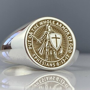 May include: A silver ring with a detailed circular emblem. The emblem features a knight with a sword and shield, surrounded by the text "PUT ON THE WHOLE ARMOR OF GOD" and "EPHESIANS 6:13-17". The ring has a polished, reflective surface.