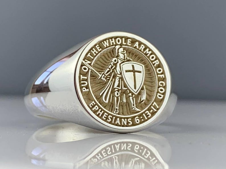 May include: A silver ring with a detailed circular emblem. The emblem features a knight with a sword and shield, surrounded by the text "PUT ON THE WHOLE ARMOR OF GOD" and "EPHESIANS 6:13-17". The ring has a polished, reflective surface.