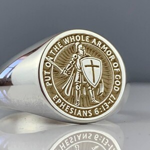 May include: Silver ring with a detailed circular emblem. The emblem features a knight with a sword and shield, surrounded by the text "PUT ON THE WHOLE ARMOR OF GOD" and "EPHESIANS 6:13-17".