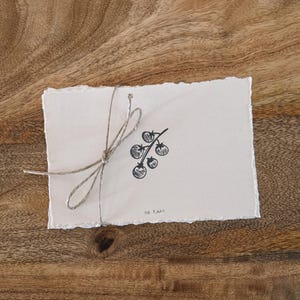 May include: A handmade, off-white card with a black illustration of tomatoes on a vine, tied with twine. The card has a deckled edge and the words "no rain" are written at the bottom. The card rests on a wooden surface.