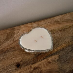 May include: A heart-shaped candle in a clear glass container. The candle has a creamy white wax and two wicks. The container has a decorative, textured design. The candle sits on a wooden surface.