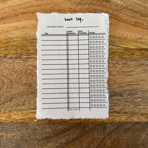 May include: A handmade book log on textured paper with a deckled edge. The black text includes fields for title, start and finish dates, and a rating system. The words "book log" are at the top. The paper rests on a wooden surface.