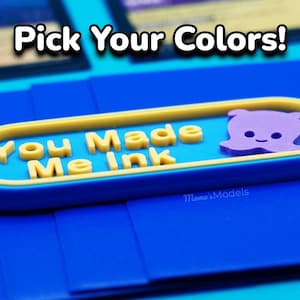 May include: A blue and yellow 3D printed sign that says "You Made Me Ink" with a purple cartoon octopus on the right side. The sign is on a blue surface.
