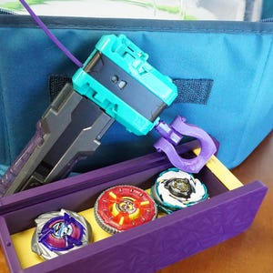 The Slip Grip in purple attached to the teal rip cord launcher next to the purple garage door style deckbox with 3 beyblades inside