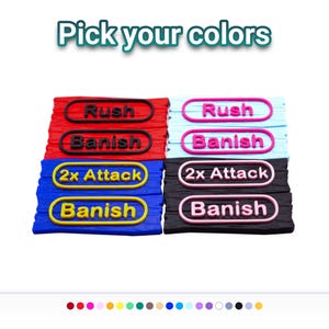 May include: A collection of rectangular game tokens with wood-grain texture, featuring raised text in oval frames. The text reads Rush, Banish, and 2x Attack. The tokens are available in red, blue, light blue, and black, with contrasting colored lettering.