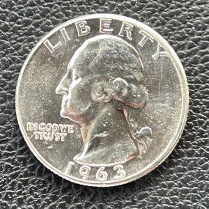 Può includere: Una moneta da un quarto di dollaro americano in argento con l'anno 1963 inciso sul fondo. La moneta presenta un profilo di George Washington con le parole "LIBERTY" sopra e "IN GOD WE TRUST" sotto.