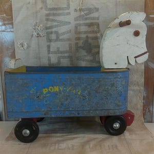 Peut inclure: Un chariot jouet en métal bleu vintage avec une tête de cheval en bois blanc. Le chariot a des roues noires avec des accents rouges et les mots "Pony-Vale" en jaune. Le fond est un sac en toile de jute.