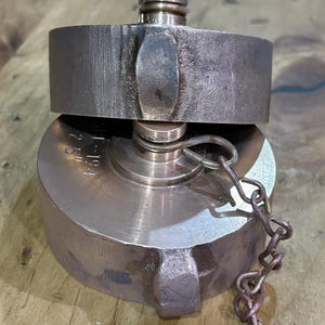 May include: A bronze-colored fire hose coupling with a chain attached. The coupling has a circular design with a raised rim and a threaded connection. The chain is made of small, linked metal pieces.