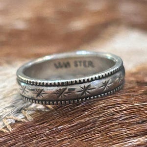 May include: A silver ring with a detailed design. The ring features a band with a repeating star pattern and a textured edge. The inscription "MM STER" is visible on the inside of the ring.
