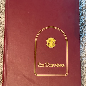 May include: A maroon-coloured hardcover book with gold lettering and design. The cover features the text "La Cumbre" and the year "1962" in a gold, stylised font within a gold arched frame. The book is closed.