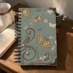 May include: A spiral-bound journal with a teal cover featuring butterfly and floral designs. The butterflies have yellow and brown details. The journal has metal corner protectors and a brown ribbon bookmark. The journal is open on a wooden surface.