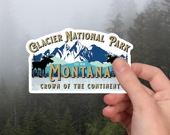 Peut inclure: Un autocollant avec les mots "Glacier National Park Montana", une chaîne de montagnes et des silhouettes d'élans. L'autocollant est bleu, blanc et beige. Le texte "Crown of the Continent" est en bas.