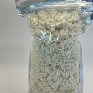May include: A clear, resealable plastic pouch filled with a granular, off-white substance. The contents appear as small, irregular, crumbly pieces with a matte texture, packed tightly within the upright, silver-trimmed storage bag.