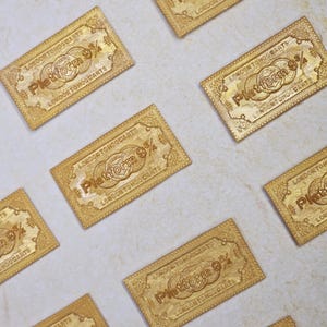May include: Gold-colored rectangular tickets with the text "Platform 9 3/4" and "London to Hogwarts" are arranged on a light-colored surface. The tickets have a vintage, ornate design.