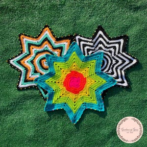 May include: Three colorful, star-shaped crochet pieces with beaded edges rest on green turf. The stars feature vibrant patterns in teal, orange, black, white, yellow, and red. The image includes a circular logo with text.