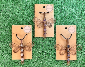 May include: Three handcrafted dragonfly pendants with copper wire bodies and gemstone accents. Each dragonfly is displayed on a brown card with a black cord. The gemstones are purple, white, and blue.