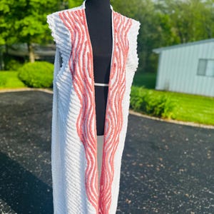 May include: A white sleeveless cardigan with a long, flowing design. The cardigan has a pink wavy pattern on the front and back.