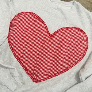 May include: A light gray sweatshirt with a large red and white gingham heart applique. The heart is outlined with a red beaded trim. The sweatshirt has long sleeves and a crew neck.