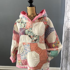 May include: A patchwork hoodie in a variety of floral and geometric patterns. The hoodie features a pink-lined hood and cuffs. The quilt-style jacket is displayed on a mannequin. The color palette includes pink, white, red, and blue.