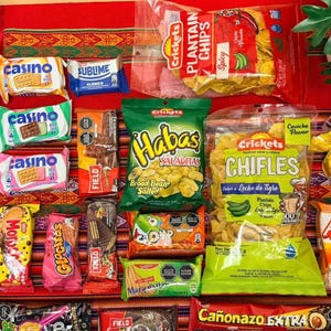 May include: A collection of various snack food packages including Crickets plantain chips, Habas broad beans, Chifles, Ajinomen instant noodles, San Jose cookies, Casino sandwich cookies, Sublime chocolate, and assorted candy bars on a red patterned surface.