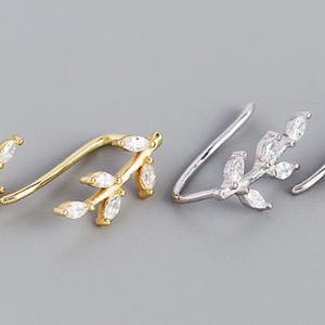 May include: A pair of ear cuffs with a leaf design. The ear cuffs are made of silver and gold metal and are adorned with clear cubic zirconia stones.