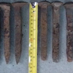 May include: A collection of 16 rusty metal railway spikes, some with square heads and some with round heads, arranged in a row on a wooden surface. The spikes are various lengths, ranging from 7.6 to 12.7 cm.