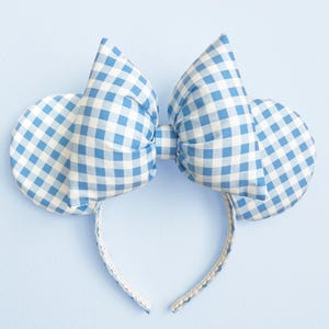 May include: A blue and white gingham patterned headband with large bow and circular ears. The headband is white with a delicate lace trim. The bow and ears are made of a light blue and white checkered fabric. The background is a solid light blue.