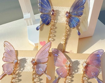 Puede incluir: Cuatro pares de pendientes con forma de mariposa en tonos azules, rosas y morados. Cada pendiente presenta alas translúcidas, un centro enjoyado y una cadena colgante con cuentas de cristal. Los pendientes se exhiben en tarjetas con el texto "Blooming Versailles".