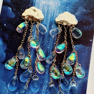 Puede incluir: Pendientes colgantes con tachuelas doradas en forma de nube. Cuentas transparentes iridiscentes en forma de lágrima cuelgan de delicadas cadenas doradas, creando un efecto de lluvia. Los pendientes están sobre un fondo azul.