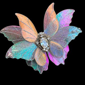 May include: A butterfly-shaped brooch featuring layered, translucent wings in iridescent shades of pink, orange, blue, and purple with silver glitter veins. The center is adorned with a clear oval-cut crystal surrounded by small, round clear stones.