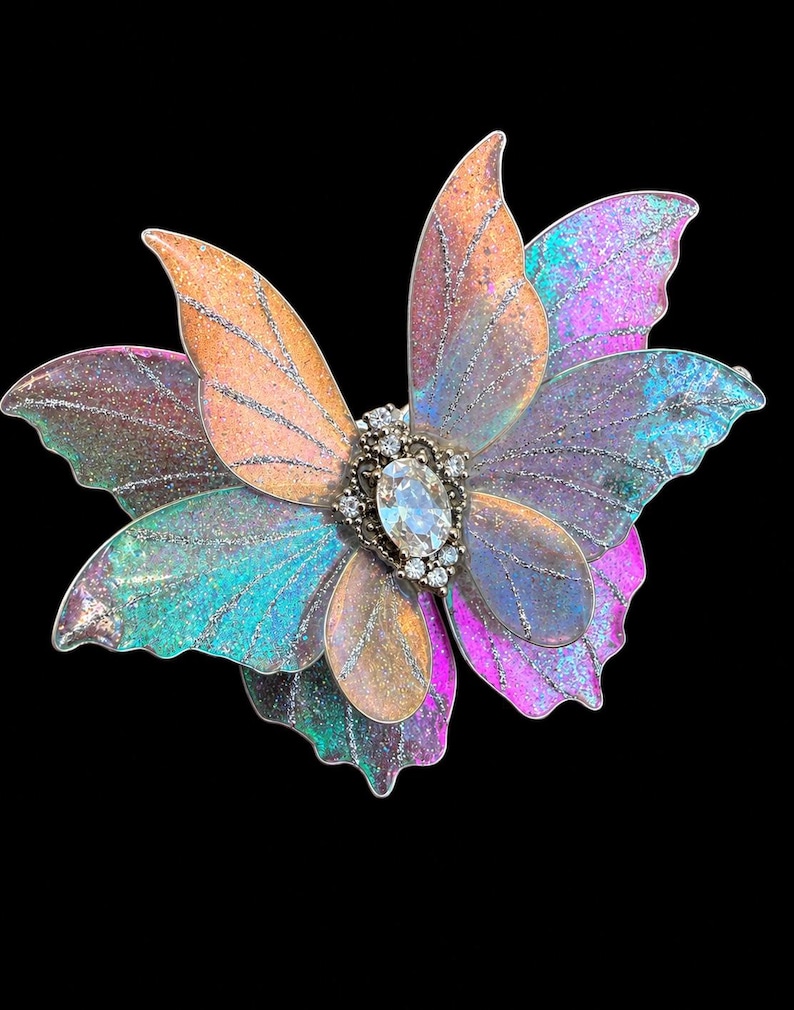 May include: A butterfly-shaped brooch featuring layered, translucent wings in iridescent shades of pink, orange, blue, and purple with silver glitter veins. The center is adorned with a clear oval-cut crystal surrounded by small, round clear stones.