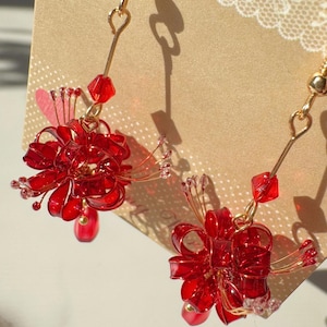 May include: A pair of red floral earrings with gold-colored hooks. Each earring features a cluster of red, translucent petals and small red beads. The earrings are designed to dangle and catch the light.