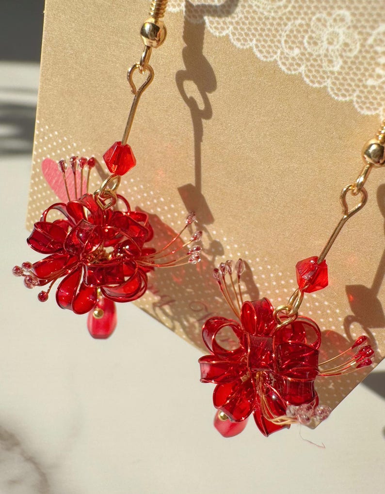 May include: A pair of red floral earrings with gold-colored hooks. Each earring features a cluster of red, translucent petals and small red beads. The earrings are designed to dangle and catch the light.