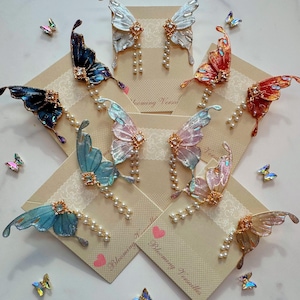 Puede incluir: Un surtido de pendientes con forma de mariposa en varios colores, incluyendo azul, rosa y naranja. Cada pendiente presenta una joya decorativa y una hilera de perlas. Los pendientes se exhiben en peque&ntilde;as tarjetas con el texto "Blooming Versailles".