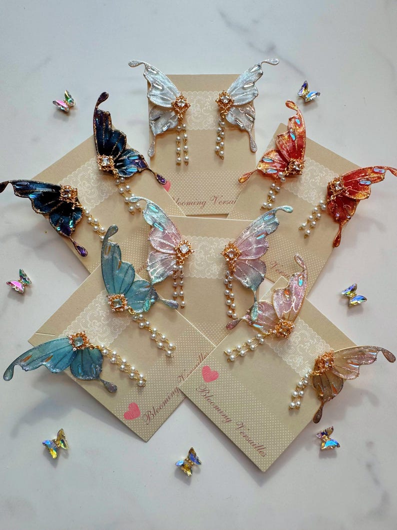 Puede incluir: Un surtido de pendientes con forma de mariposa en varios colores, incluyendo azul, rosa y naranja. Cada pendiente presenta una joya decorativa y una hilera de perlas. Los pendientes se exhiben en peque&ntilde;as tarjetas con el texto "Blooming Versailles".