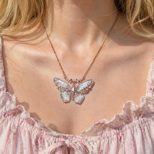 May include: A gold-toned metal necklace featuring a large, intricate butterfly pendant. The butterfly wings have a translucent, iridescent finish with delicate filigree detailing and small clear crystals embedded along the edges and center.