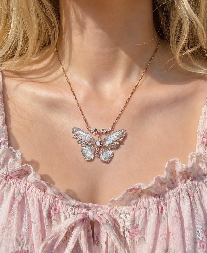May include: A gold-toned metal necklace featuring a large, intricate butterfly pendant. The butterfly wings have a translucent, iridescent finish with delicate filigree detailing and small clear crystals embedded along the edges and center.