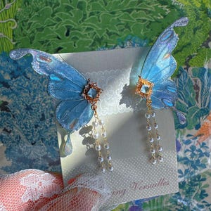Puede incluir: Un par de pendientes de alas de mariposa azules con detalles brillantes. Cada pendiente presenta una joya central y perlas de imitaci&oacute;n colgantes. Los pendientes se colocan sobre un fondo con el texto "Versailles".