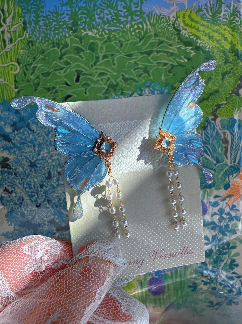 Puede incluir: Un par de pendientes de alas de mariposa azules con detalles brillantes. Cada pendiente presenta una joya central y perlas de imitaci&oacute;n colgantes. Los pendientes se colocan sobre un fondo con el texto "Versailles".