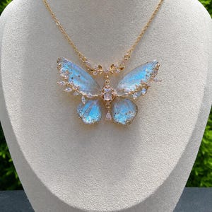 May include: A gold-toned necklace featuring a large butterfly pendant with iridescent blue and white wings. The butterfly is accented with clear, sparkling crystals along the edges and a central rectangular stone, suspended from a delicate gold chain.
