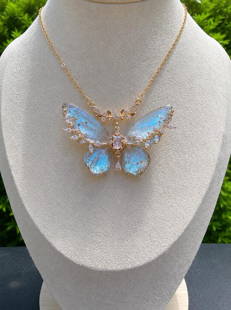May include: A gold-toned necklace featuring a large butterfly pendant with iridescent blue and white wings. The butterfly is accented with clear, sparkling crystals along the edges and a central rectangular stone, suspended from a delicate gold chain.
