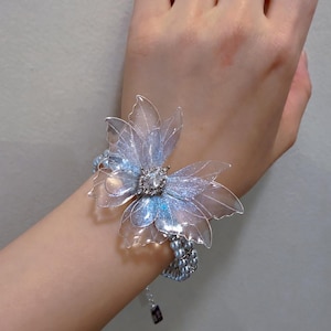 May include: A bracelet featuring a large, translucent, iridescent butterfly-shaped ornament with shimmering blue and purple accents. The center of the butterfly is adorned with a clear, faceted crystal. The band consists of multiple strands of small, round, light blue pearls.