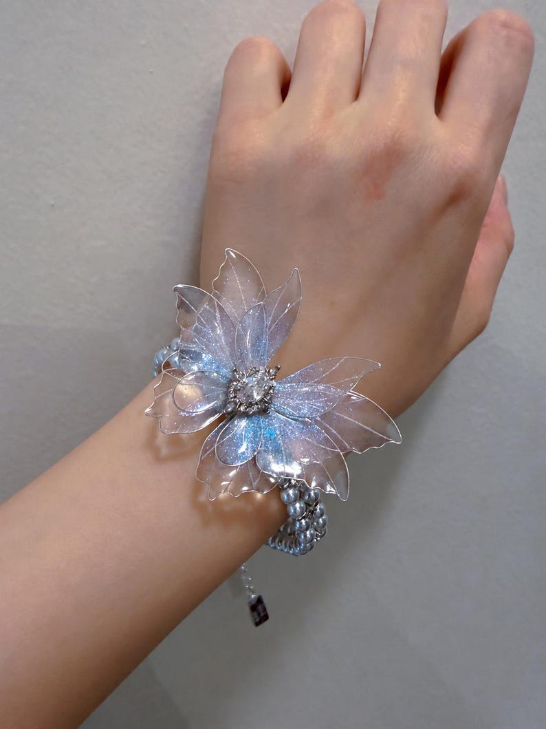 May include: A bracelet featuring a large, translucent, iridescent butterfly-shaped ornament with shimmering blue and purple accents. The center of the butterfly is adorned with a clear, faceted crystal. The band consists of multiple strands of small, round, light blue pearls.