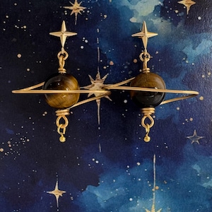 以下が含まれることがあります： 虎目石の球体と金の星チャームが付いたゴールドのイヤリング。球体は、惑星のリングシステムのような金のリングの中に吊り下げられています。