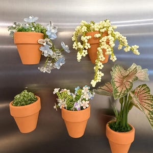 May include: Five miniature terracotta-colored flower pots functioning as magnets. The pots contain various decorative plants made of beads, including blue flowers, white berries on green vines, green foliage, multicolored small flowers, and a plant with pink and green leaves.