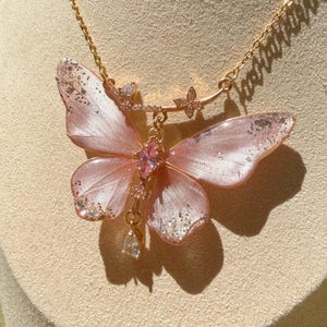 May include: A gold-tone chain necklace featuring a large, translucent pink butterfly pendant with silver glitter accents on the wing tips. The butterfly center is adorned with a marquise-cut pink crystal, and a clear teardrop crystal hangs below.